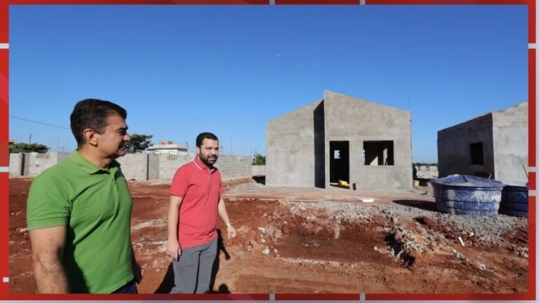 Two men in polo shirts walk across a red-soil construction site with unfinished concrete houses and blue sky in the background, showing open doorways and windows.