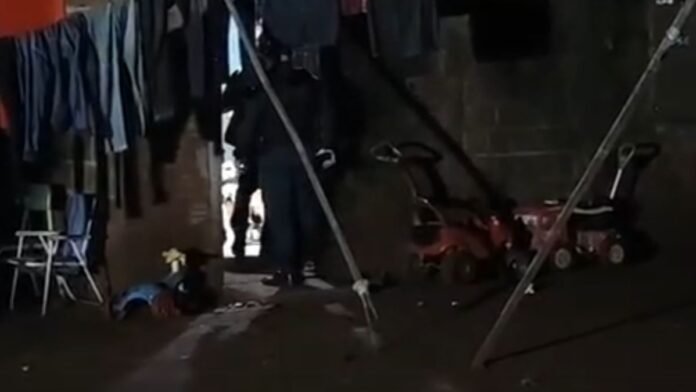 Person standing in a dim, cluttered room with clothing hanging and lawn equipment around (lawnmowers, chairs).