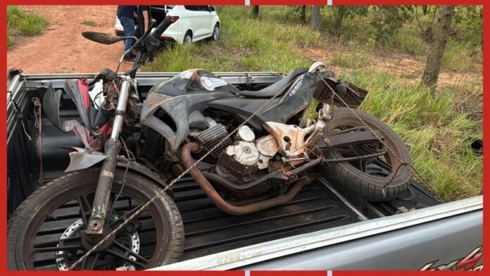 Damaged motorcycle tied down in the bed of a pickup truck on a dirt road, showing extensive rust and missing parts.