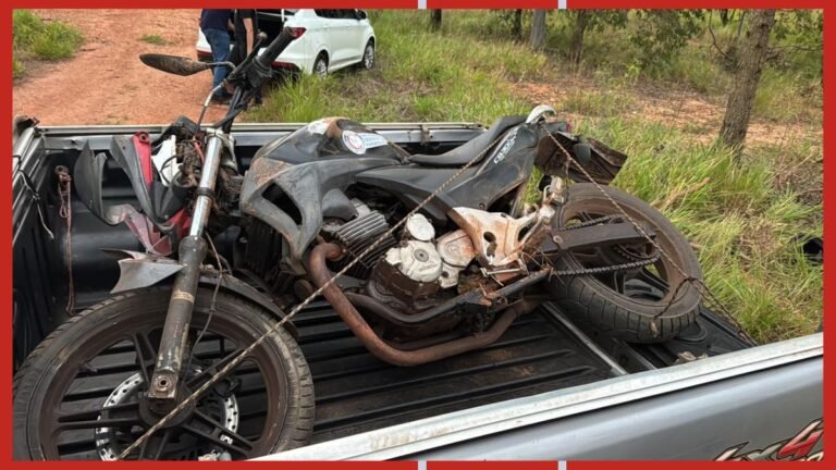 Damaged motorcycle tied down in the bed of a pickup truck on a dirt road, showing extensive rust and missing parts.