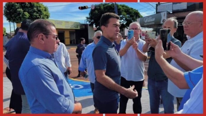 Man in a dark polo shirt stands center while supporters surround him, taking photos with smartphones outdoors.
