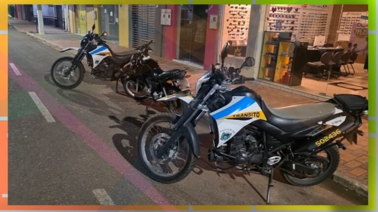 Two police motorcycles labeled Transito parked on a city street at night near storefronts and a sidewalk streaked with colorful pavement markings.