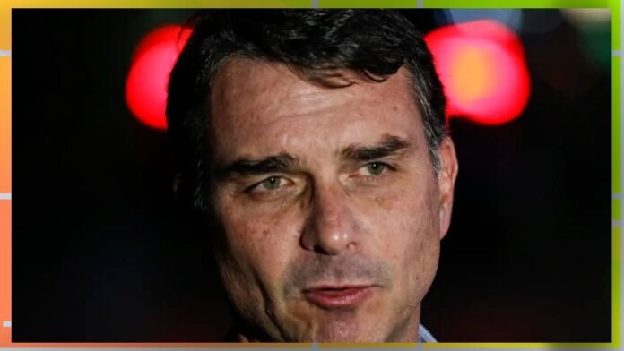 Close-up of a middle-aged man with short dark hair and slight stubble against a dark background with red lights in the distance.