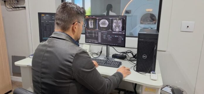 Radiology technician reviews MRI/CT images on dual computer monitors beside the MRI suite window.