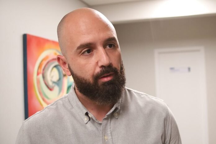 Bearded, bald man in a light gray button-down shirt speaks, standing in a hallway with abstract art in the background.