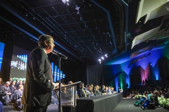 A man in a suit speaks into a microphone at a clear podium on a lit stage, with a long panel of people seated behind him and a large audience in the background.