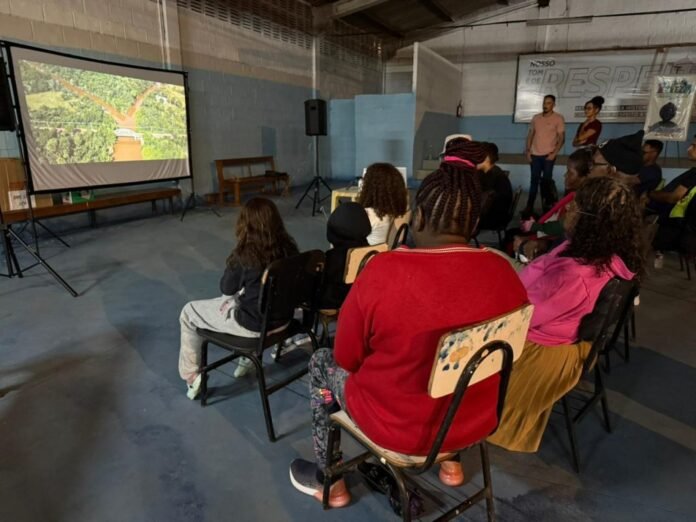 Group of people seated in a hall watching a projected nature image on a screen side-by-side with a bench and speaker stands nearby.