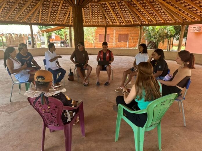 Group of people seated in a circle under a thatched outdoor shelter, engaged in a discussion.