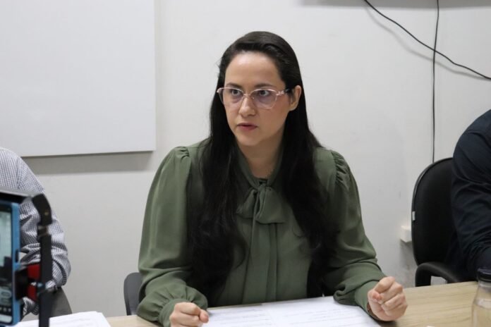 Woman in a green blouse with a bow at the neck sits at a conference table, speaking and looking to the side, with papers in front of her.