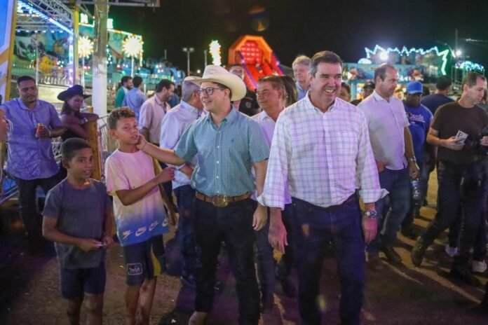 A lively nighttime carnival scene with adults and children walking among colorful rides and lights; a man in a light plaid shirt smiles at the front.