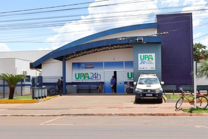 A medical clinic with a blue curved roof and a sign that reads UPA 24h, Unidade de Pronto Atendimento, in front of a parking area and a white pickup truck.