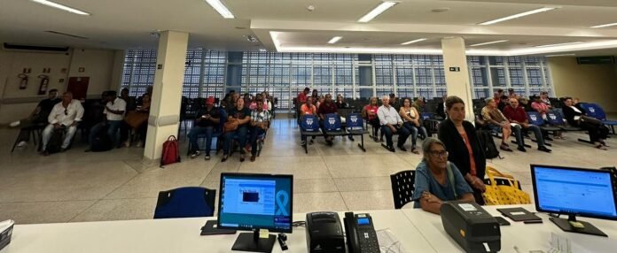People seated in a large waiting area, facing the camera, with desks and computer monitors in the foreground.