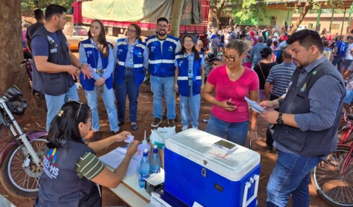 Group of volunteers in blue vests stand in a circle at an outdoor community event while a woman sits at a table filling out forms next to a blue cooler and water bottles.