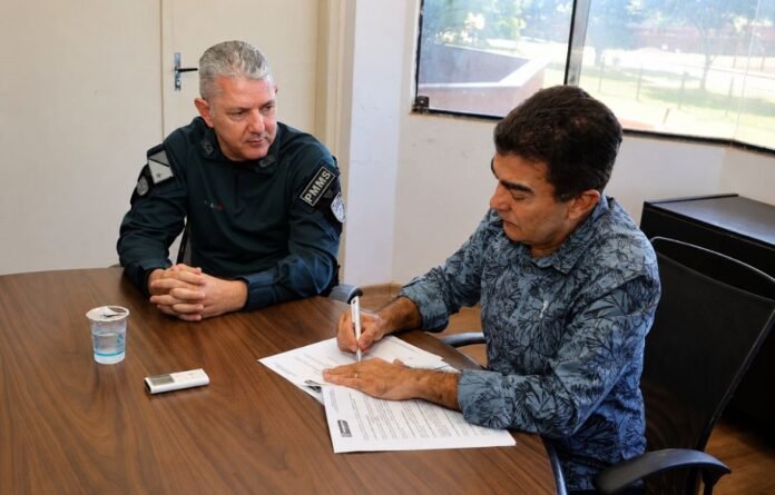 Two men sit at a wooden table; one in a dark uniform watches as the other signs documents nearby.