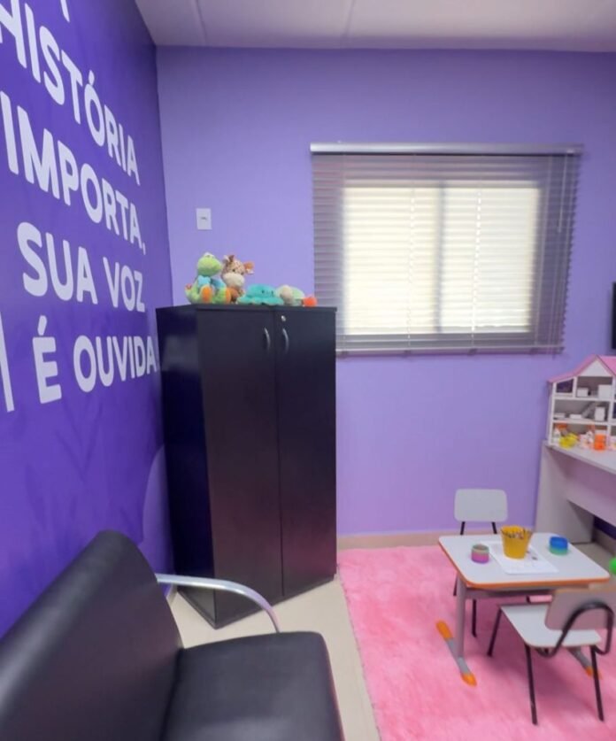 Playroom with purple walls, large white Portuguese text, a black cabinet topped with plush toys, a pink rug, and a small table with chairs.