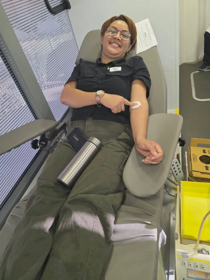 Woman donating blood, seated in a medical chair, arm extended with a cotton ball and bandage after needle insertions, smiling at camera.