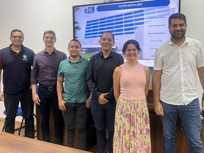 Six professionals stand side by side in an office, smiling, with a hospital presentation slide on a screen behind them.