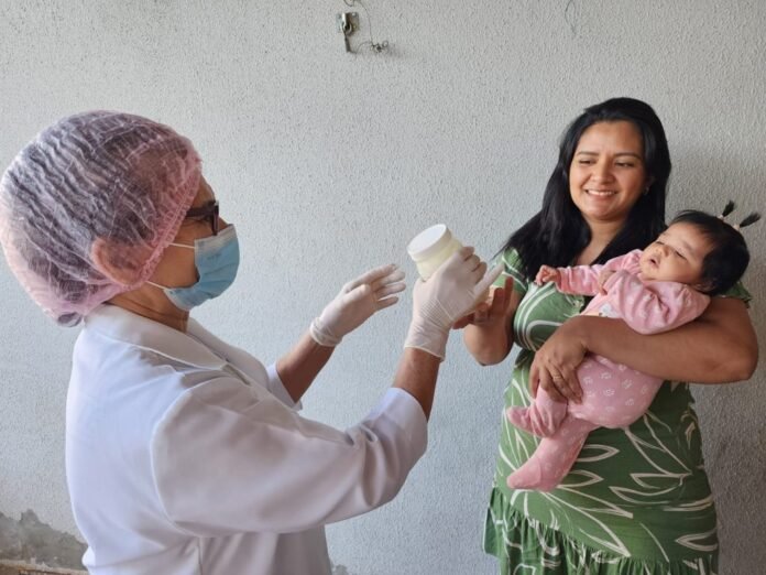 Healthcare worker in a mask, hairnet, and gloves offers a bottle to a mother holding her baby indoors.