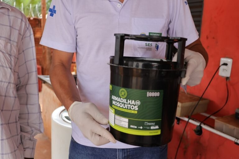 Health worker wearing gloves holds a black mosquito trap bucket labeled 'Armadilha Mosquitos' in a health outreach setting.