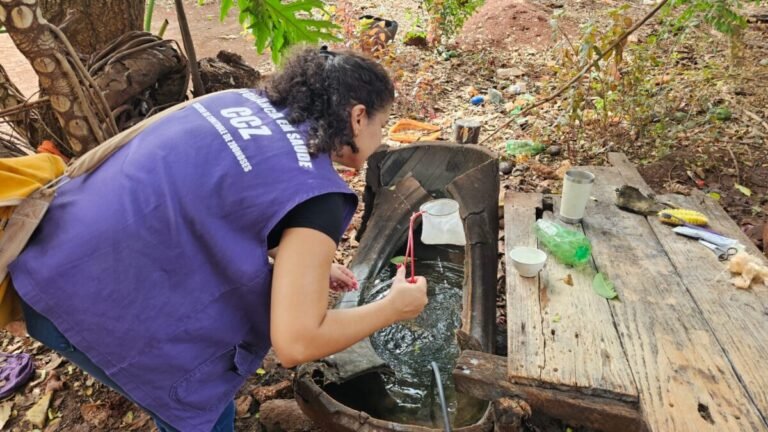 Field worker in a purple vest collecting a water sample from a barrel outdoors, using a red sampling tool near a weathered wooden table with cups and tools nearby.