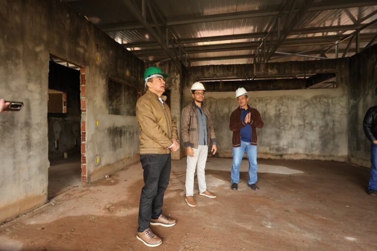 Three men in hard hats stand inside an unfinished building under a metal roof, surveying the space.