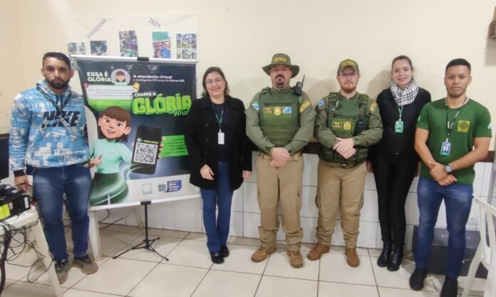 Six people stand indoors beside a colorful poster about Glória, including two uniformed officers; everyone wears lanyards. The poster features a cartoon child and a QR code.