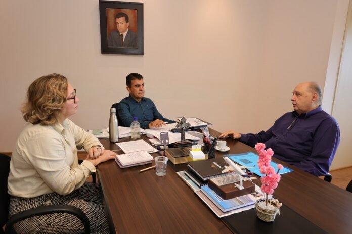 Three professionals sit around a wooden conference table in an office, discussing documents and notes laid out before them, with a portrait hanging on the wall.