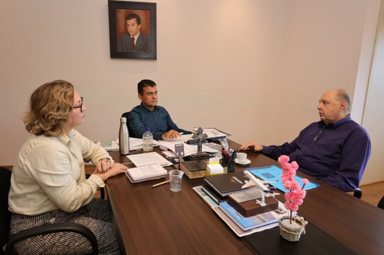 Three professionals sit around a wooden conference table in an office, discussing documents and notes laid out before them, with a portrait hanging on the wall.
