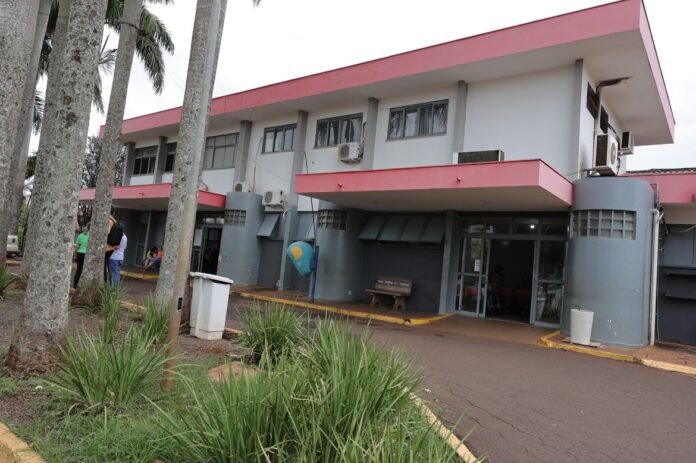 Exterior view of a gray two-story building with pink trim and a glass entrance, palm trees in the foreground and people near the doorway.