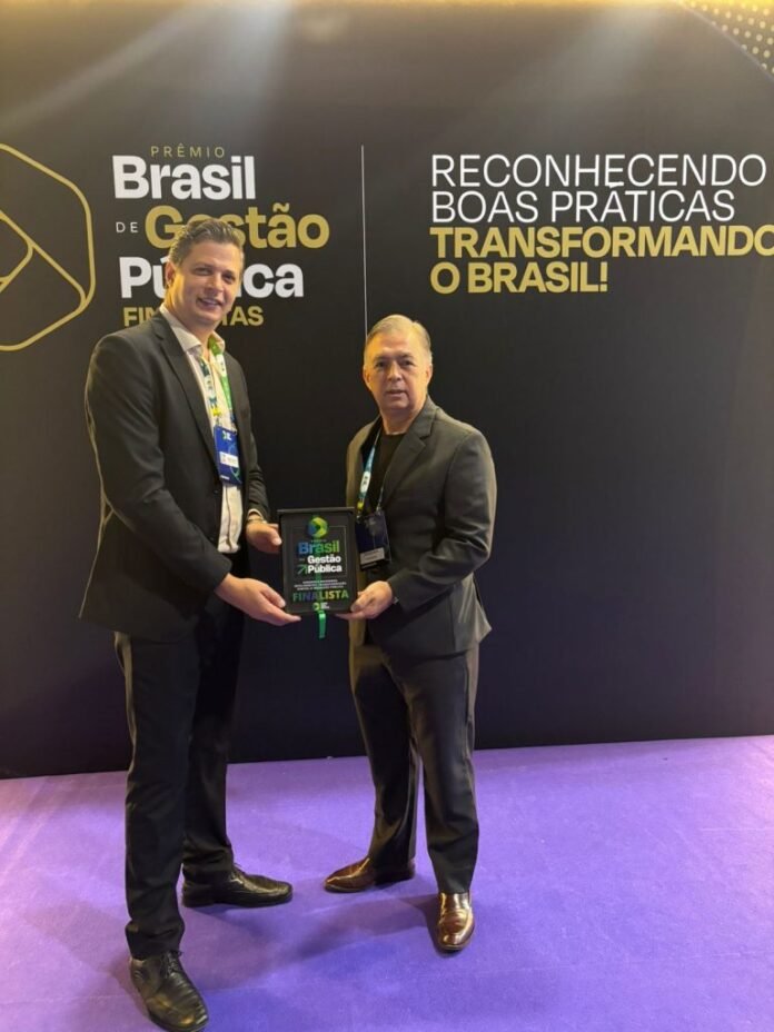 Two men in suits stand on a purple carpet as one hands a plaque to the other at an awards event with Portuguese signage in the background.