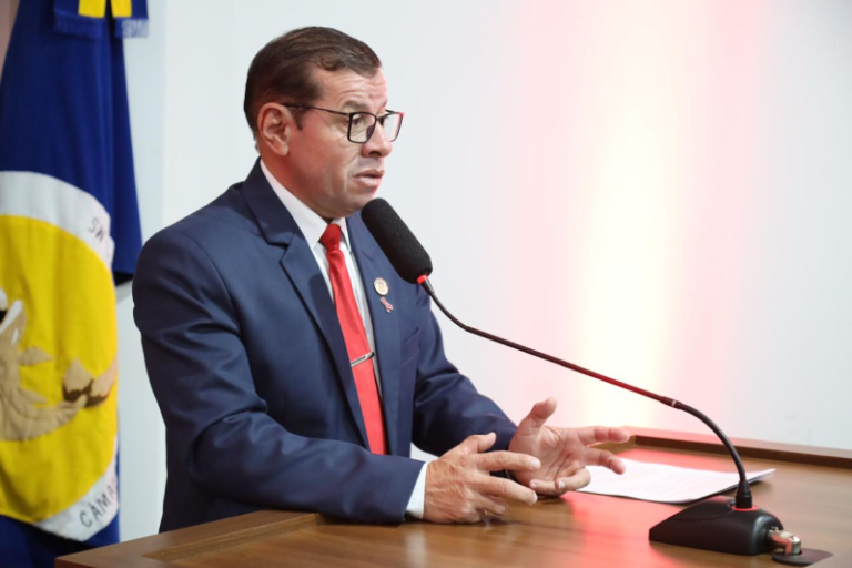 Man in a navy suit and red tie speaks into a microphone at a lectern, flag in the background.