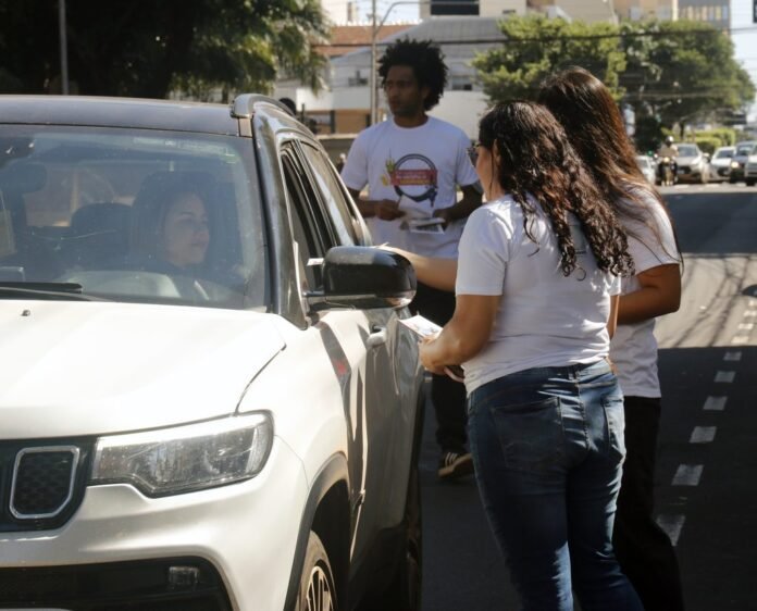 Volunteer in a white shirt hands a paper to a driver in a white SUV on a sunny street; other volunteers stand nearby.