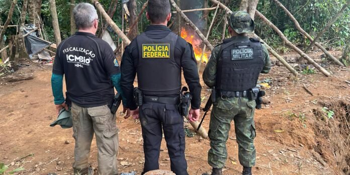 Three uniformed officers with backs to the camera stand around a small campfire in a wooded area.