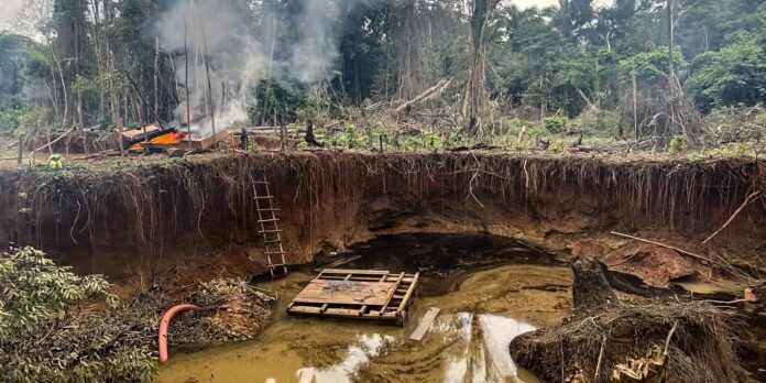 Smoke rises from a burned wooden shelter on a muddy riverbank, with a wooden ladder leaning from the bank.