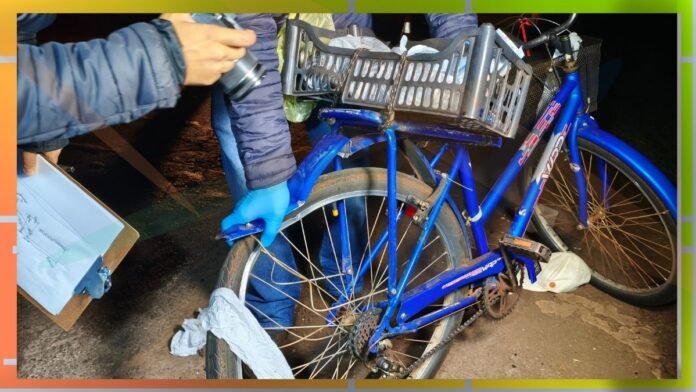 Blue bicycle with a front basket being inspected at night; gloved hand holding a camera near the front wheel.