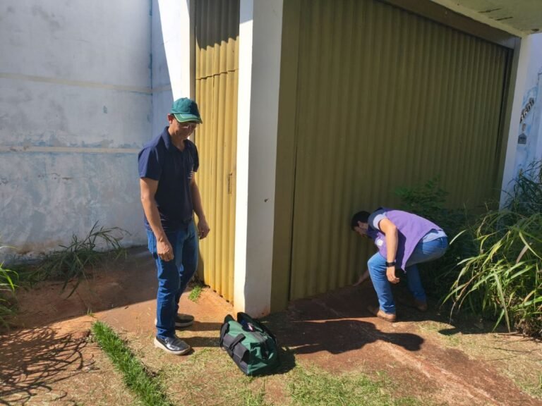 Two men outdoors near a green metal door; one stands watching while the other crouches to inspect the ground, with a green and black bag nearby.