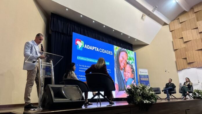 Speaker at a podium on stage with a large blue presentation screen that reads 'ADAPTA CIDADES' and shows a smiling woman holding a child in the background.