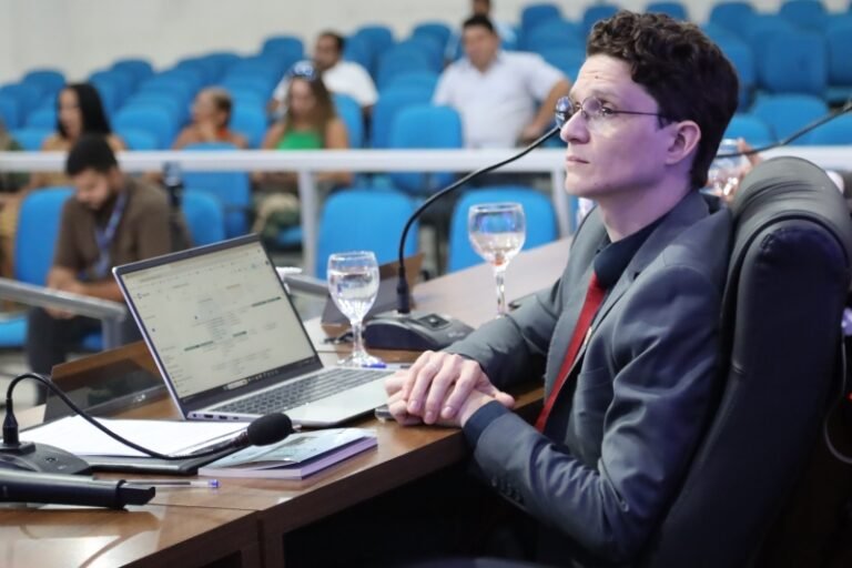 Panelist in a gray suit and red tie sits at a desk with a laptop, microphone, and glasses in a conference hall with blue seats in the background.