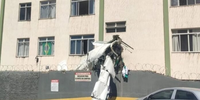 A mangled metal sculpture resembling an airplane wreck atop a high wall with barbed wire, in front of a beige apartment building; Brazilian flag in a window.