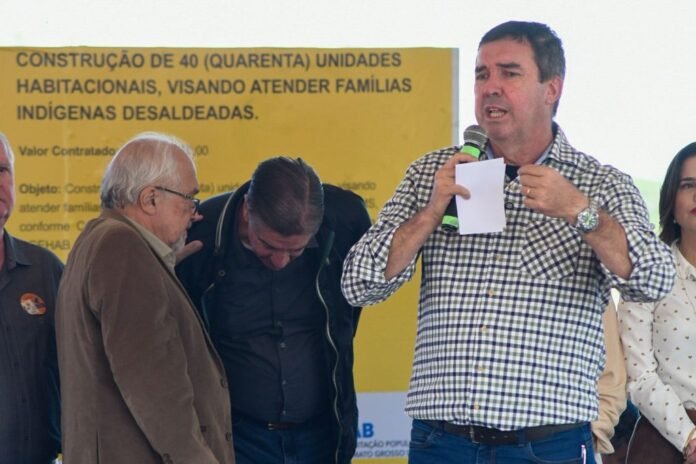 Man in a checkered shirt speaks into a mic and holds a note, standing at a public event with a yellow banner about building 40 housing units for indigenous families.