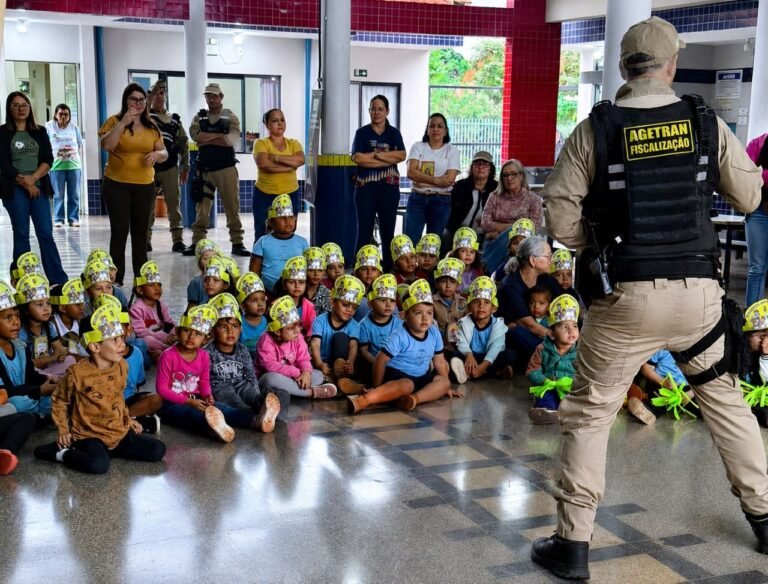 Group of children wearing yellow crown hats sit on the floor as a security officer addresses them; adults stand in the background.