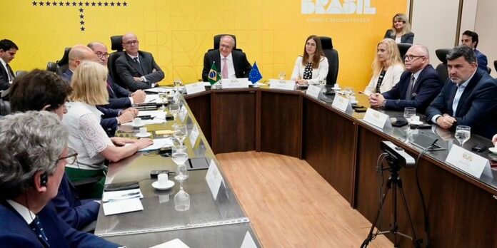 Officials sit around a U-shaped conference table in a bright yellow room for a formal meeting, with Brazilian and EU flags on the table nearby.