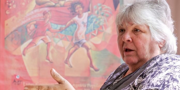 Portrait of an elderly woman with short gray hair speaking, with a colorful mural of dancers in the background.