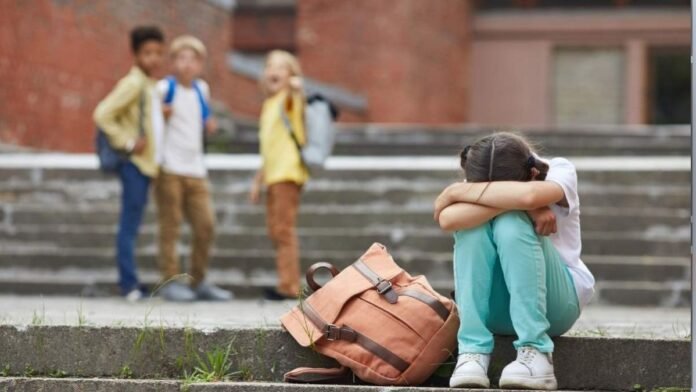 A girl sits on the steps with her arms folded over her knees, a peach backpack beside her, while three classmates stand nearby.