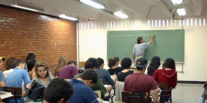 Instructor writes on a green chalkboard at the front of a classroom while students sit at desks and take notes.
