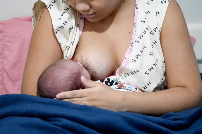 Mother breastfeeding her newborn, cradling the baby at her chest as the infant nurses at the breast with a blue blanket nearby.