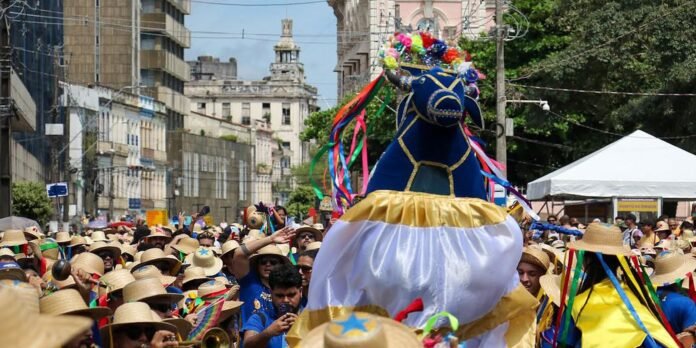 Colorful street parade with a large blue float shaped like a horse wearing a golden ribboned dress, surrounded by a crowd in straw hats on a city street.