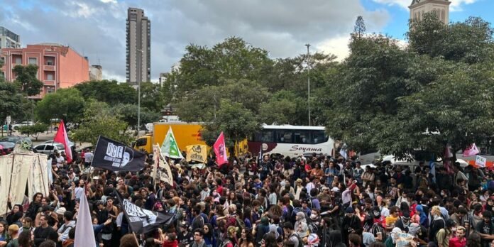 Large diverse crowd gathered for a protest in an urban square with colorful flags and banners.