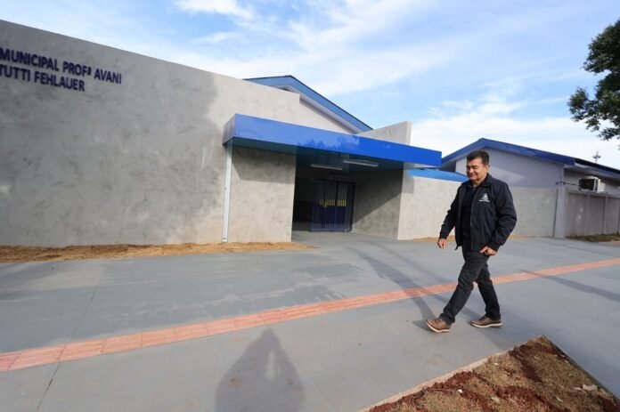 Man in a dark jacket and brown shoes walks along a concrete path outside a gray municipal building with a blue awning under a clear sky.