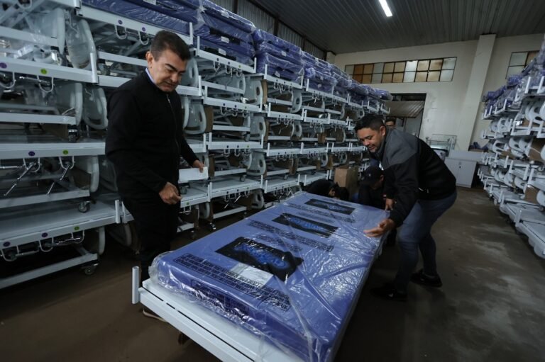 Workers move a blue plastic–wrapped hospital mattress on a wheeled bed frame in a storage warehouse with rows of stacked beds along the walls.